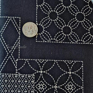 May include: A black fabric featuring white stitched geometric patterns in a sashiko style. The textile displays repeating circular and diamond motifs arranged in rectangular sections. A silver-colored coin is placed on the fabric for scale.