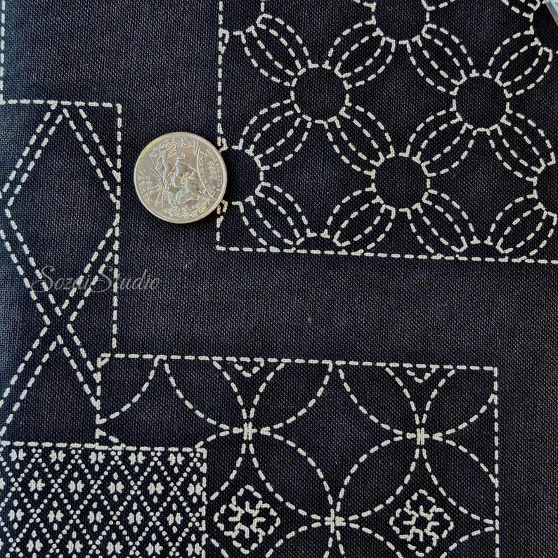 May include: A black fabric featuring white stitched geometric patterns in a sashiko style. The textile displays repeating circular and diamond motifs arranged in rectangular sections. A silver-colored coin is placed on the fabric for scale.