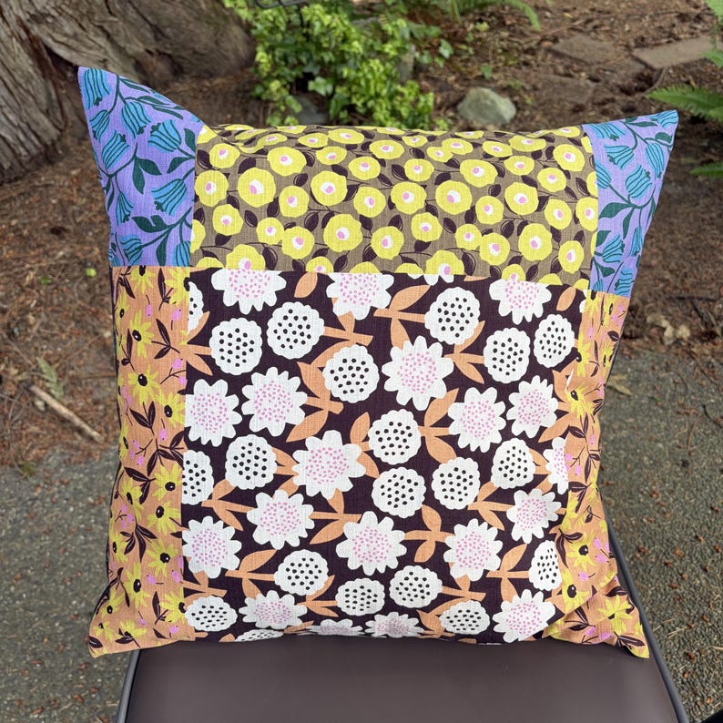 Peut inclure: Un coussin carr&eacute; pr&eacute;sentant un motif patchwork compos&eacute; de divers tissus &agrave; fleurs. Le panneau central affiche des fleurs blanches et roses sur fond marron fonc&eacute;, entour&eacute; de motifs floraux jaunes et de fleurs en cloche bleues aux coins.