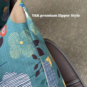 May include: A teal throw pillow cover featuring a floral pattern with yellow, orange, and dark brown elements, including striped and checkered shapes. The corner shows a white YKK premium Zipper Style closure integrated into the fabric seam.