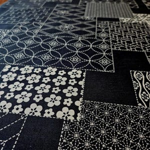 May include: A black fabric featuring a white patchwork design composed of various geometric and floral patterns. The textile displays repeating motifs including circles, stars, and blossoms arranged in a grid-like layout across the surface.