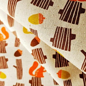 Pu&ograve; includere: Primo piano di un tessuto beige con un motivo ripetuto di tronchi d'albero marroni, scoiattoli arancioni e ghiande gialle e marroni. Il tessuto &egrave; piegato, mostrando la trama e il design. Il tema generale &egrave; il bosco.