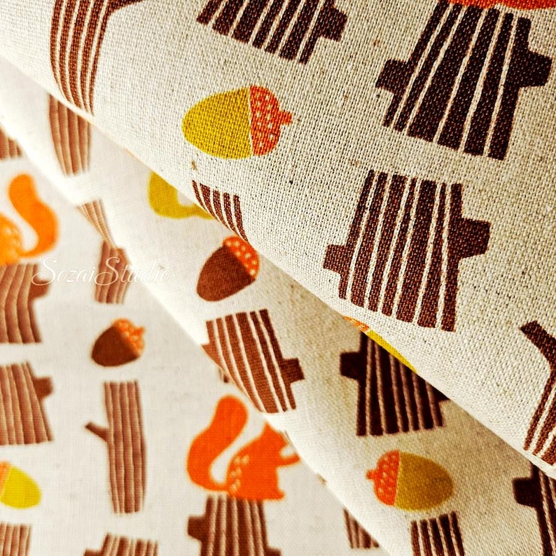 Pu&ograve; includere: Primo piano di un tessuto beige con un motivo ripetuto di tronchi d'albero marroni, scoiattoli arancioni e ghiande gialle e marroni. Il tessuto &egrave; piegato, mostrando la trama e il design. Il tema generale &egrave; il bosco.