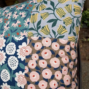 K&ouml;nnte beinhalten: Ein quadratisches Zierkissen mit einem Patchwork-Design aus vier verschiedenen floralen Stoffmustern. Die Abschnitte zeigen hellrosa G&auml;nsebl&uuml;mchen auf Petrol, gelbe Tulpen auf Hellblau, wei&szlig;e Blumen auf Dunkelblau und rosa Bl&uuml;ten auf Braun.