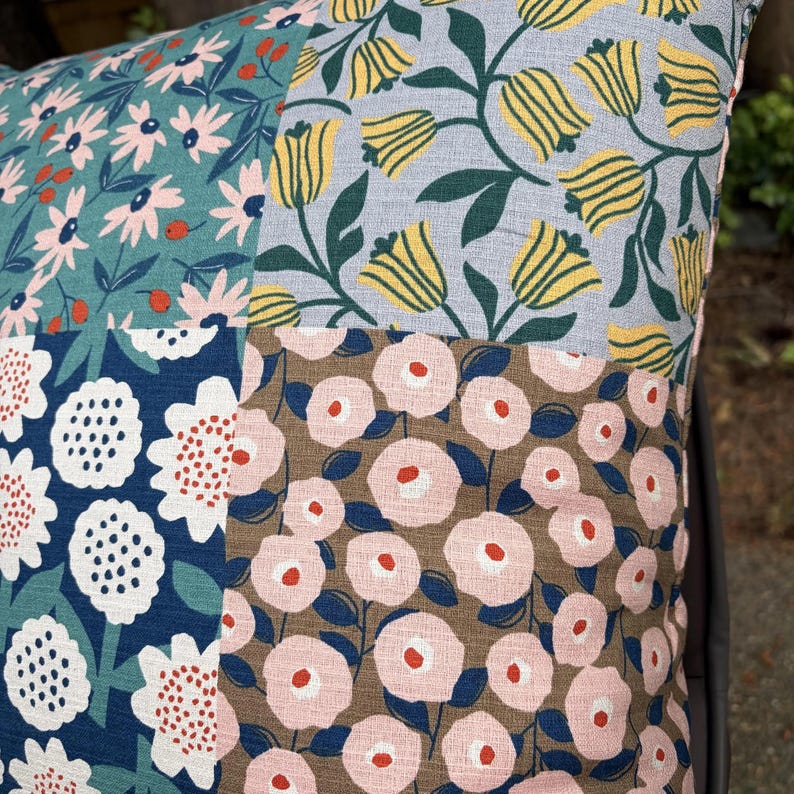 K&ouml;nnte beinhalten: Ein quadratisches Zierkissen mit einem Patchwork-Design aus vier verschiedenen floralen Stoffmustern. Die Abschnitte zeigen hellrosa G&auml;nsebl&uuml;mchen auf Petrol, gelbe Tulpen auf Hellblau, wei&szlig;e Blumen auf Dunkelblau und rosa Bl&uuml;ten auf Braun.