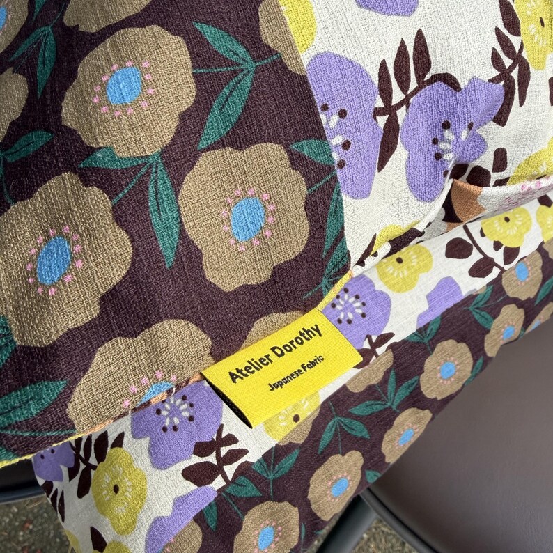 Peut inclure: Une housse de coussin en patchwork avec un motif floral compos&eacute; de fleurs marron, cr&egrave;me, violettes et jaunes. Une &eacute;tiquette en tissu jaune vif est cousue sur la couture, portant le texte noir Atelier Dorothy et Japanese Fabric.