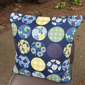 May include: A square throw pillow featuring a dark navy blue background covered in a repeating pattern of large circles. Each circle contains distinct designs including yellow gingham, floral motifs in blue, yellow, and white, and vertical stripes.