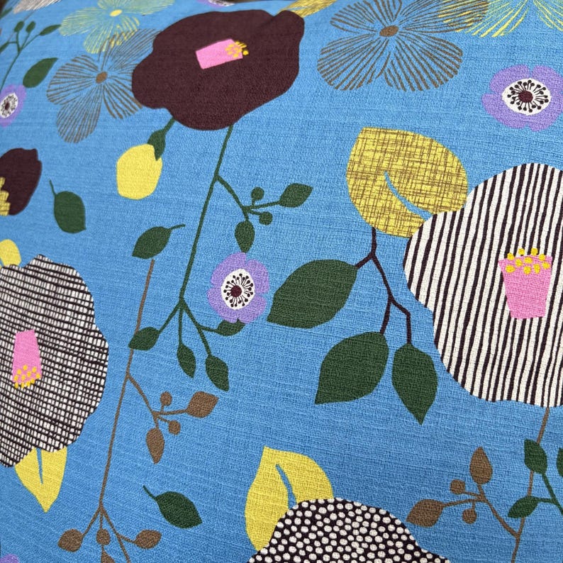 Puede incluir: Una tela azul brillante con un patr&oacute;n floral de flores de color marr&oacute;n oscuro, rayas blancas y negras, y flores moradas. El dise&ntilde;o incluye hojas amarillas, follaje verde oscuro y tallos marrones finos sobre la superficie texturizada.
