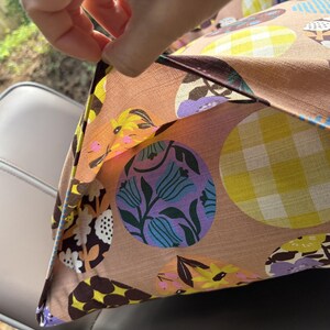 K&ouml;nnte beinhalten: Ein Kissenbezug aus Stoff mit beigefarbener Grundfl&auml;che und einem wiederkehrenden Muster aus gro&szlig;en Kreisen. Die Kreise enthalten verschiedene Designs, darunter gelb-wei&szlig;es Vichy-Karo, violette Tulpen mit gr&uuml;nen Bl&auml;ttern und dunkelbraune Blumenmotive.