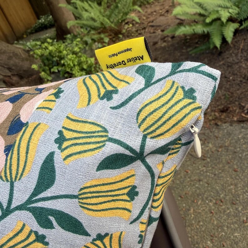 K&ouml;nnte beinhalten: Ein hellblauer Kissenbezug mit einem gewebten Muster aus gelben Tulpen mit dunkelgr&uuml;nen Stielen und Bl&auml;ttern. Am oberen Rand befindet sich ein kleines gelbes Stoffetikett mit der Aufschrift Atelier Dorothy Japanese Fabric und ein wei&szlig;er Rei&szlig;verschluss.