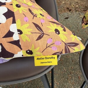 Peut inclure: Un coussin carr&eacute; en tissu beige avec un motif r&eacute;p&eacute;titif de fleurs jaunes, de feuilles brun fonc&eacute; et de petites baies roses. Une &eacute;tiquette en tissu jaune est fix&eacute;e sur le c&ocirc;t&eacute;, portant le texte Atelier Dorothy Japanese Fabric.