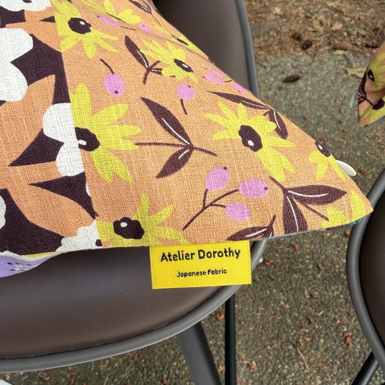 Peut inclure: Un coussin carr&eacute; en tissu beige avec un motif r&eacute;p&eacute;titif de fleurs jaunes, de feuilles brun fonc&eacute; et de petites baies roses. Une &eacute;tiquette en tissu jaune est fix&eacute;e sur le c&ocirc;t&eacute;, portant le texte Atelier Dorothy Japanese Fabric.
