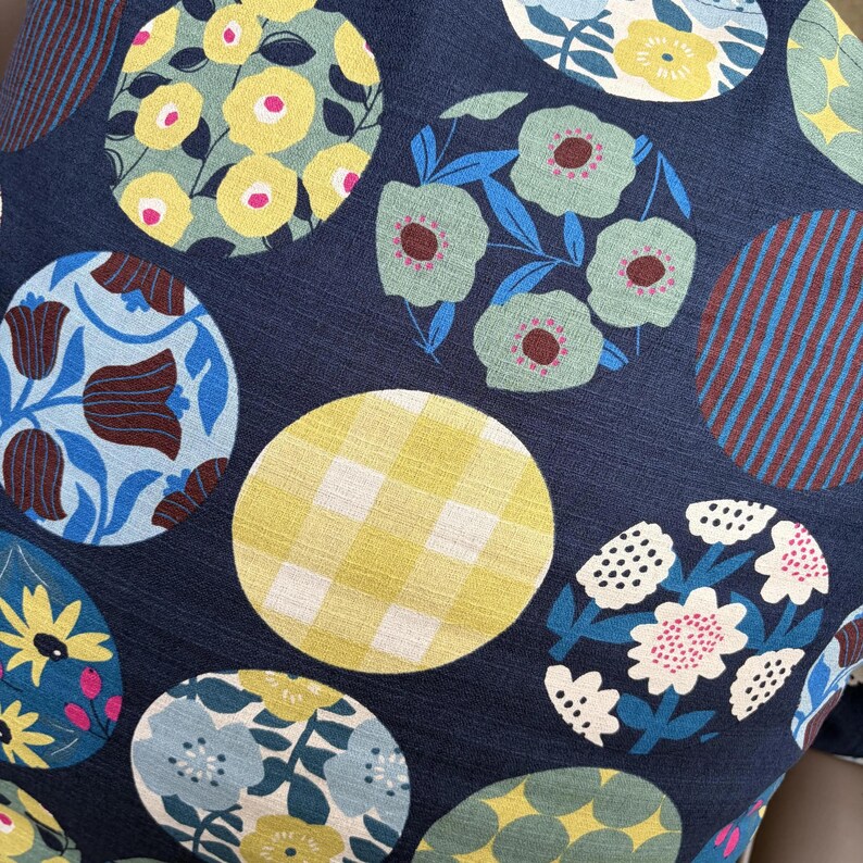 May include: A dark navy blue fabric featuring a repeating pattern of circular motifs. The circles contain various designs including yellow flowers, light green blossoms, brown and blue tulips, a yellow gingham check, and white floral clusters.