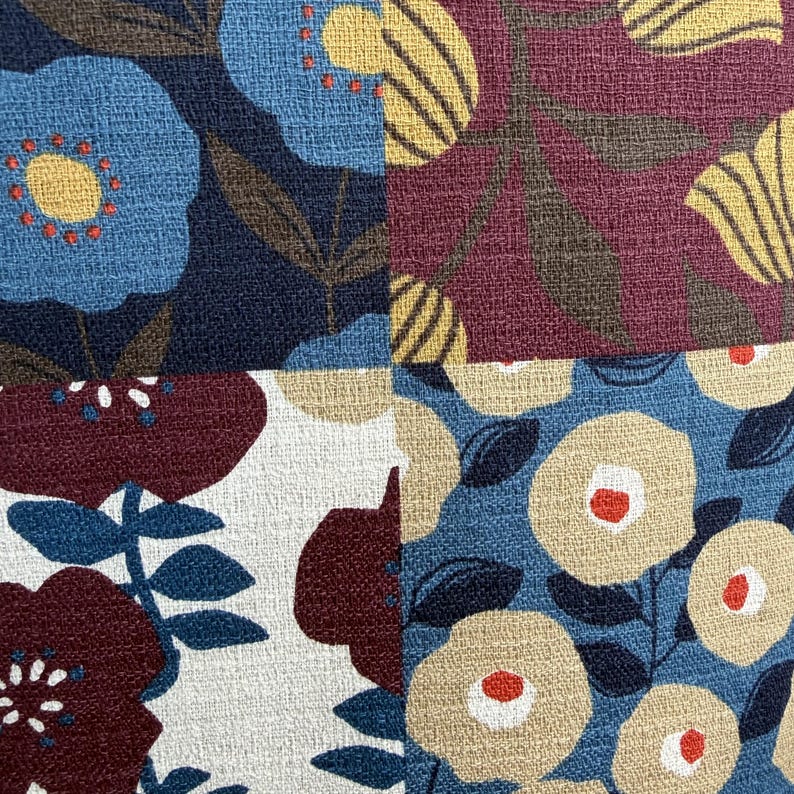 May include: A textile fabric featuring a four-quadrant patchwork design. The pattern displays stylized floral motifs in shades of navy blue, burgundy, cream, and yellow, with orange accents on the flower centers, all set against a textured weave.