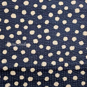 May include: Dark blue fabric with a textured grid pattern and scattered white polka dots. The dots have a slightly irregular, brushstroke-like appearance. The fabric has a subtle sheen, suggesting a potential use in apparel or home decor.