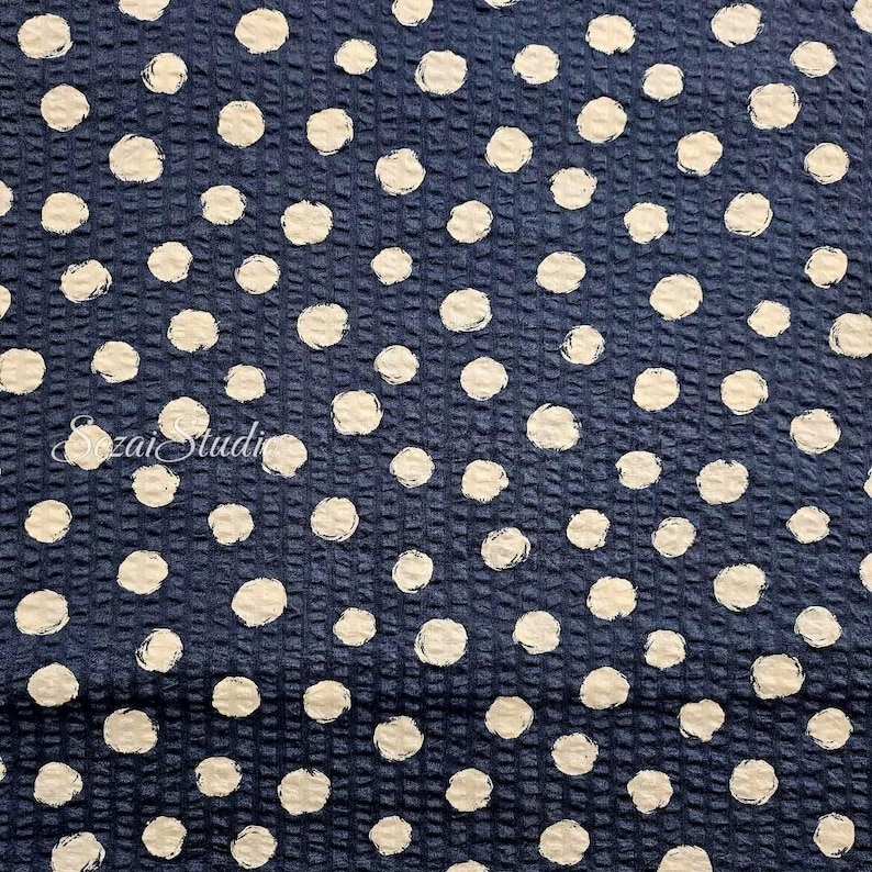 May include: Dark blue fabric with a textured grid pattern and scattered white polka dots. The dots have a slightly irregular, brushstroke-like appearance. The fabric has a subtle sheen, suggesting a potential use in apparel or home decor.