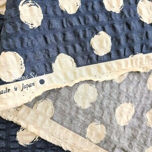 May include: Close-up of blue fabric with large white polka dots. A strip of off-white fabric with the text "Made by Japan" is visible. The fabric has a textured, waffle-like appearance.