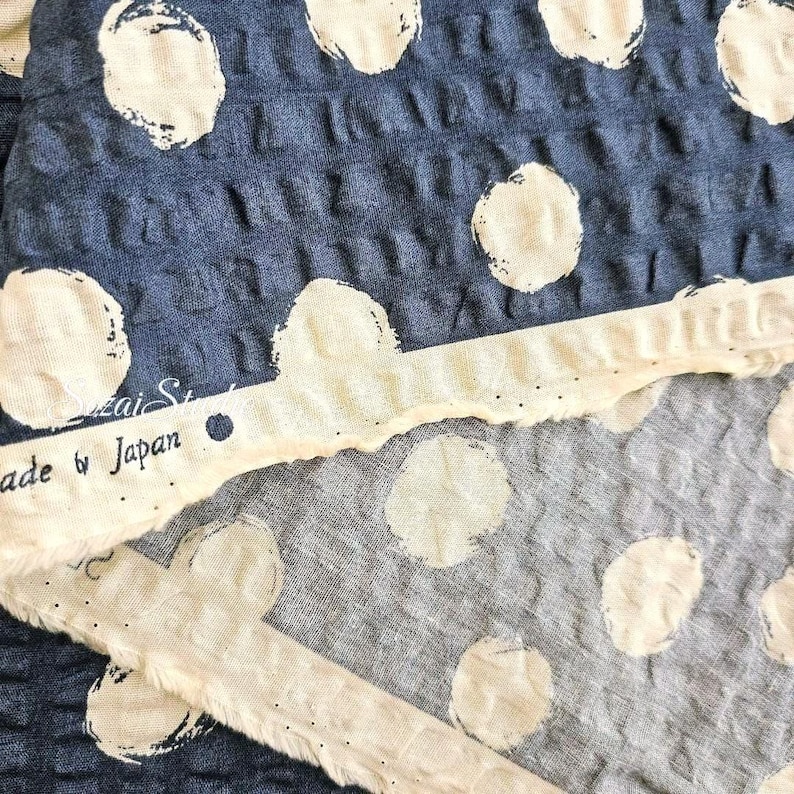 May include: Close-up of blue fabric with large white polka dots. A strip of off-white fabric with the text "Made by Japan" is visible. The fabric has a textured, waffle-like appearance.