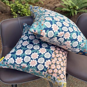 K&ouml;nnte beinhalten: Zwei quadratische Dekokissen mit Patchwork-Design und Blumenmustern in Marineblau, Petrol, Hellrosa und Wei&szlig;. Der Stoff zeigt verschiedene botanische Motive, darunter stilisierte Blumen, Bl&auml;tter und kleine kreisf&ouml;rmige Punkte.