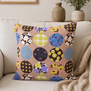 May include: A square throw pillow with a tan background featuring a repeating pattern of circles. Each circle contains different designs including yellow gingham, purple and yellow flowers, dark brown floral prints, and blue vertical stripes.
