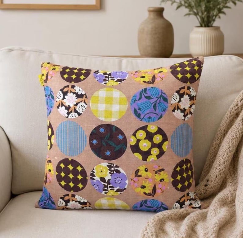 K&ouml;nnte beinhalten: Ein quadratisches Dekokissen mit beigem Hintergrund und einem sich wiederholenden Kreismuster. Jeder Kreis enth&auml;lt verschiedene Designs, darunter gelbes Vichy-Karo, violette und gelbe Blumen, dunkelbraune Blumenmuster und blaue Streifen.