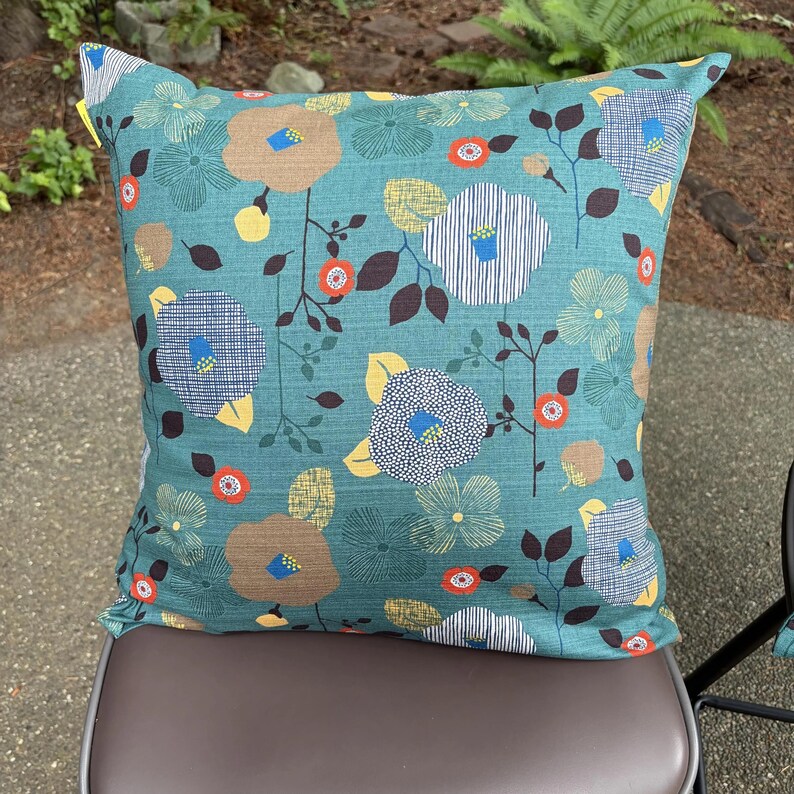 May include: A square throw pillow featuring a teal background with a repeating floral pattern. The design includes stylized flowers in shades of tan, blue, yellow, and white, accented by small orange circular blossoms and dark brown leaves.