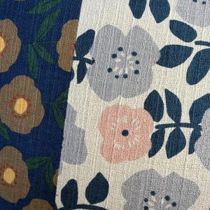 K&ouml;nnte beinhalten: Zwei St&uuml;cke strukturierten Stoffes mit Blumenmustern. Die linke Seite ist dunkelblau mit beigen Blumen und gr&uuml;nen Bl&auml;ttern. Die rechte Seite ist cremewei&szlig; mit hellblauen und zartrosa Blumen, dunkelblauen Zentren und dunkelblauen Bl&auml;ttern.