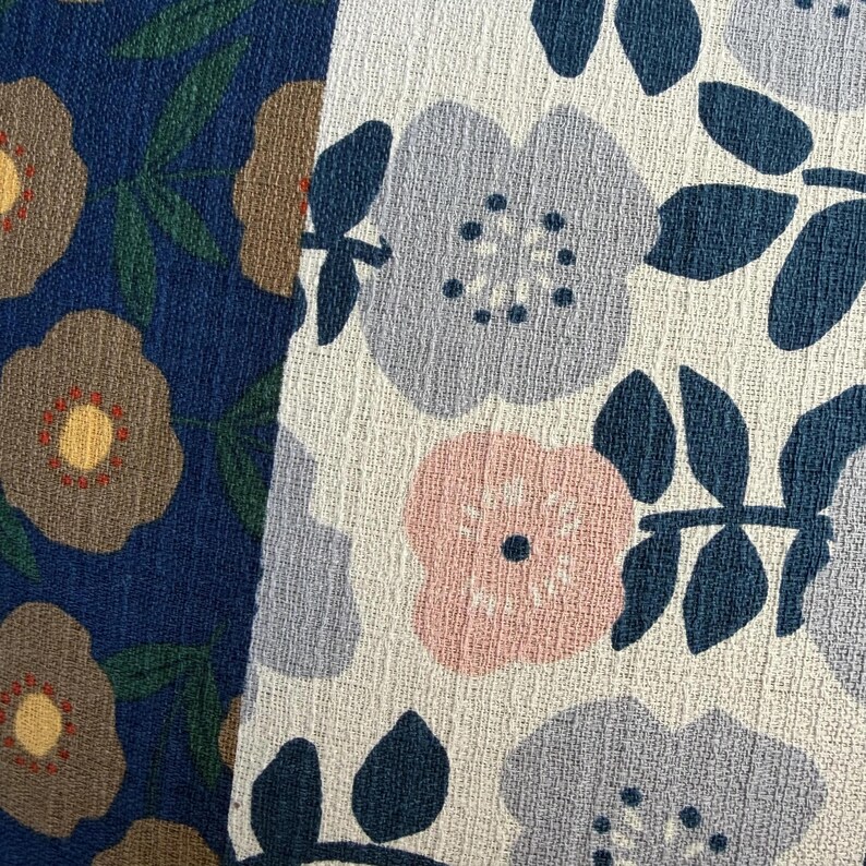 K&ouml;nnte beinhalten: Zwei St&uuml;cke strukturierten Stoffes mit Blumenmustern. Die linke Seite ist dunkelblau mit beigen Blumen und gr&uuml;nen Bl&auml;ttern. Die rechte Seite ist cremewei&szlig; mit hellblauen und zartrosa Blumen, dunkelblauen Zentren und dunkelblauen Bl&auml;ttern.