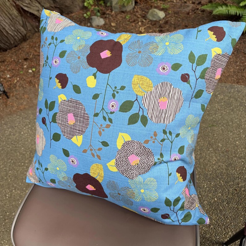 Puede incluir: Un coj&iacute;n cuadrado con fondo azul claro y un patr&oacute;n floral repetitivo. El dise&ntilde;o incluye flores de color marr&oacute;n oscuro, amarillo y azul claro con hojas y tallos verdes. Algunas flores presentan texturas a rayas o puntos con centros de color rosa.
