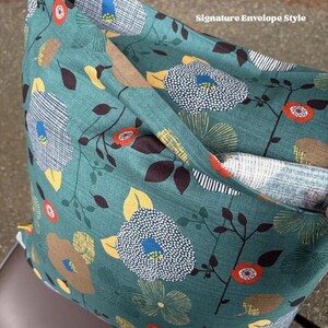 May include: A square throw pillow cover featuring a teal background with a floral pattern of white, yellow, orange, and brown flowers and leaves. The fabric has a signature envelope style closure visible on the front, with a folded flap design.