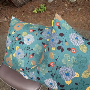 May include: Two square throw pillows featuring a teal background with a repeating floral pattern. The design includes stylized flowers in white, yellow, and brown, accented by small orange circles and dark brown leaves. A small yellow tag is visible.