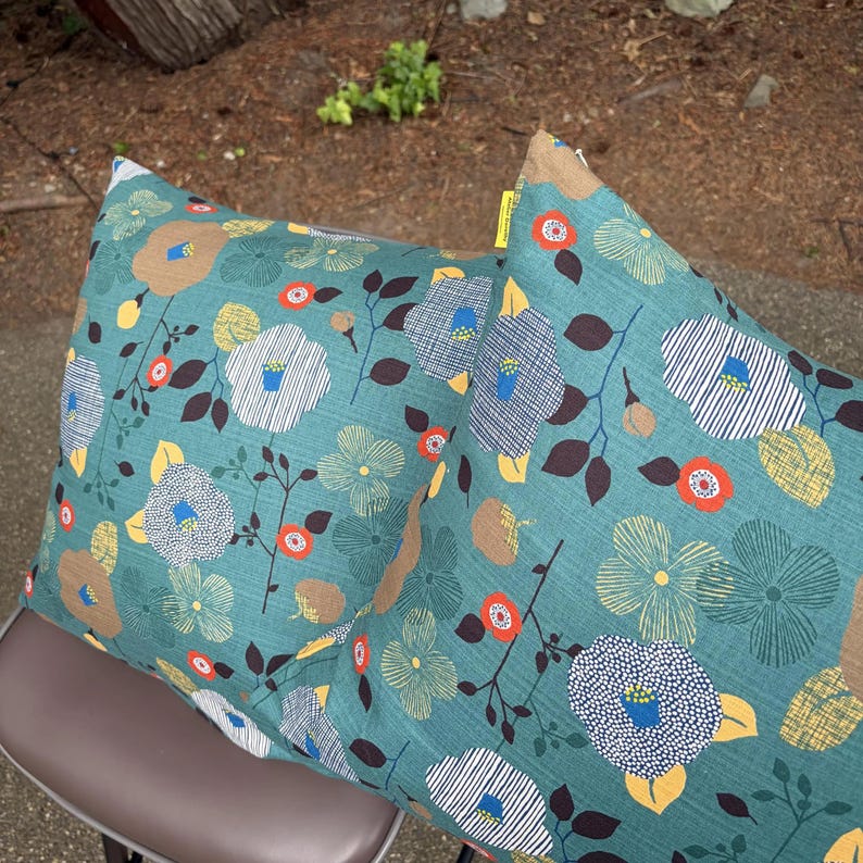 May include: Two square throw pillows featuring a teal background with a repeating floral pattern. The design includes stylized flowers in white, yellow, and brown, accented by small orange circles and dark brown leaves. A small yellow tag is visible.