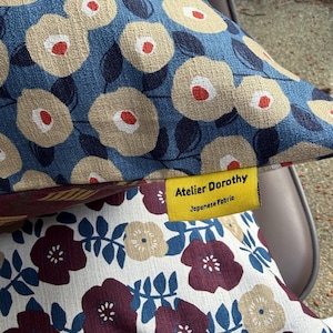 May include: A decorative pillow featuring a blue fabric with a beige and red floral pattern, layered over a white fabric with a dark red and beige floral pattern. A yellow fabric tag is attached, printed with the text Atelier Dorothy Japanese Fabric.