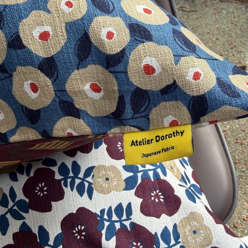 May include: A decorative pillow featuring a blue fabric with a beige and red floral pattern, layered over a white fabric with a dark red and beige floral pattern. A yellow fabric tag is attached, printed with the text Atelier Dorothy Japanese Fabric.