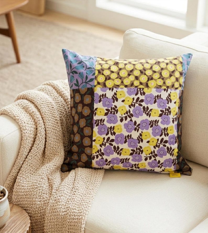 Peut inclure: Un coussin carr&eacute; pr&eacute;sentant un motif patchwork de fleurs dans des tons de jaune, violet et marron. Le coussin est compos&eacute; de quatre panneaux de tissu distincts, chacun affichant un imprim&eacute; botanique diff&eacute;rent avec des fleurs de tailles vari&eacute;es.