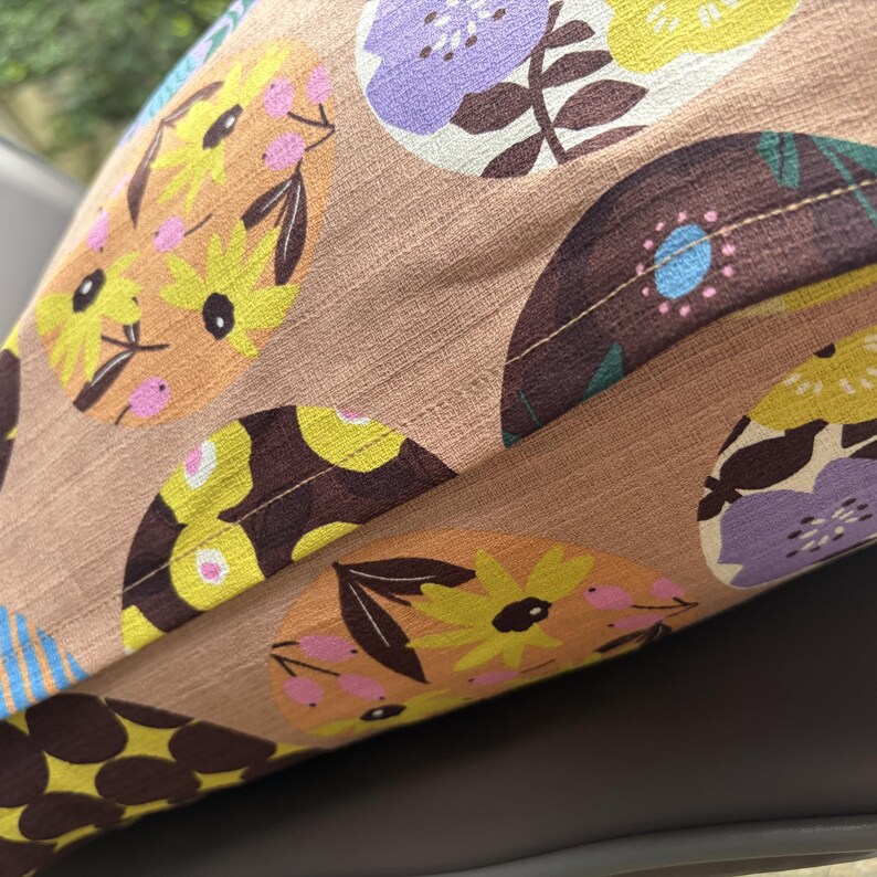 K&ouml;nnte beinhalten: Ein beigefarbener Kissenbezug aus Stoff mit einem sich wiederholenden Muster aus kreisf&ouml;rmigen Motiven. Jeder Kreis enth&auml;lt stilisierte Blumendesigns in Gelb-, Lila- und Dunkelbraunt&ouml;nen, akzentuiert durch kleine rosa Punkte und dunkelgr&uuml;ne Bl&auml;tter auf strukturiertem Grund.