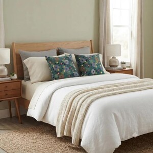 May include: A bed featuring a white duvet cover, a cream-colored knit throw blanket, and two square throw pillows with a dark teal floral pattern. The bed is styled with two solid gray pillows and two solid cream pillows against a wooden headboard.