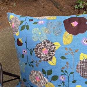 Puede incluir: Un coj&iacute;n cuadrado con un fondo azul brillante y un patr&oacute;n floral repetitivo. El dise&ntilde;o incluye flores estilizadas en tonos marr&oacute;n oscuro, amarillo, azul claro y morado, acentuadas con hojas verdes y tallos delgados de color marr&oacute;n.