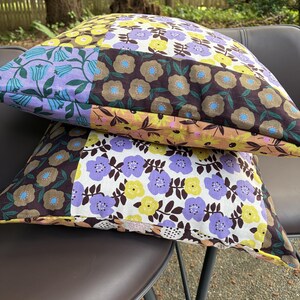 Peut inclure: Deux coussins carr&eacute;s pr&eacute;sentant un motif patchwork de fleurs. Le tissu affiche diverses sections avec des fonds marron, jaune, violet clair et blanc, d&eacute;cor&eacute;es de motifs floraux stylis&eacute;s dans des couleurs contrastantes.