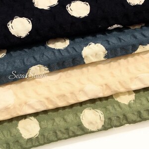 May include: Close-up of four fabric swatches in navy, teal, beige, and olive green. Each fabric has a textured surface with large, off-white polka dots. The brand name "Soza Studio" is visible on the teal fabric.