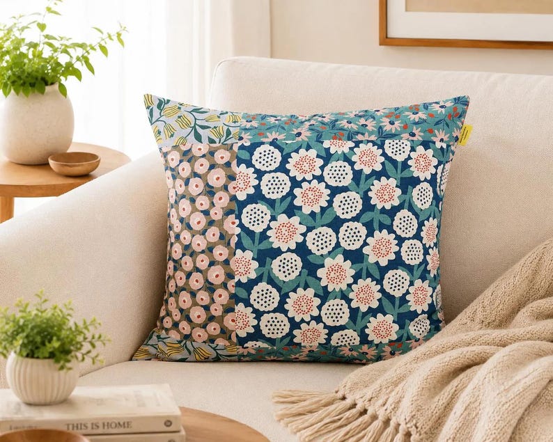 K&ouml;nnte beinhalten: Ein quadratisches Zierkissen mit Patchwork-Design und verschiedenen Blumenmustern. Der Stoff zeigt eine Mischung aus Dunkelblau, Hellrosa und Gelb mit stilisierten Blumenmotiven, darunter wei&szlig;e Bl&uuml;tenbl&auml;tter mit gepunkteten Mitten.
