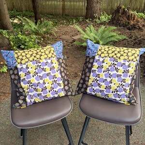 Peut inclure: Deux coussins carr&eacute;s pr&eacute;sentant un motif patchwork de fleurs dans des tons de jaune, violet et brun fonc&eacute;. Le tissu affiche des motifs floraux r&eacute;p&eacute;titifs avec des tiges et des feuilles vertes, termin&eacute;s par une petite &eacute;tiquette jaune sur le c&ocirc;t&eacute;.