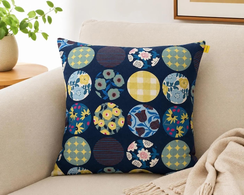 May include: A square throw pillow featuring a dark navy blue background with a repeating grid of circular patterns. Each circle displays a unique design, including yellow gingham, blue and yellow floral motifs, and geometric shapes in shades of blue, yellow, and white.
