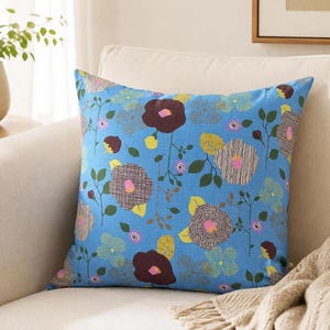 May include: A square throw pillow featuring a bright blue background with a repeating pattern of stylized flowers in shades of dark brown, yellow, light blue, and white, accented with small green leaves and thin brown stems.
