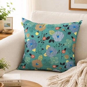 May include: A square throw pillow featuring a teal green background with a repeating pattern of stylized flowers in shades of blue, tan, yellow, and orange, accented by black stems and leaves. A small yellow tag is visible on the upper left side.