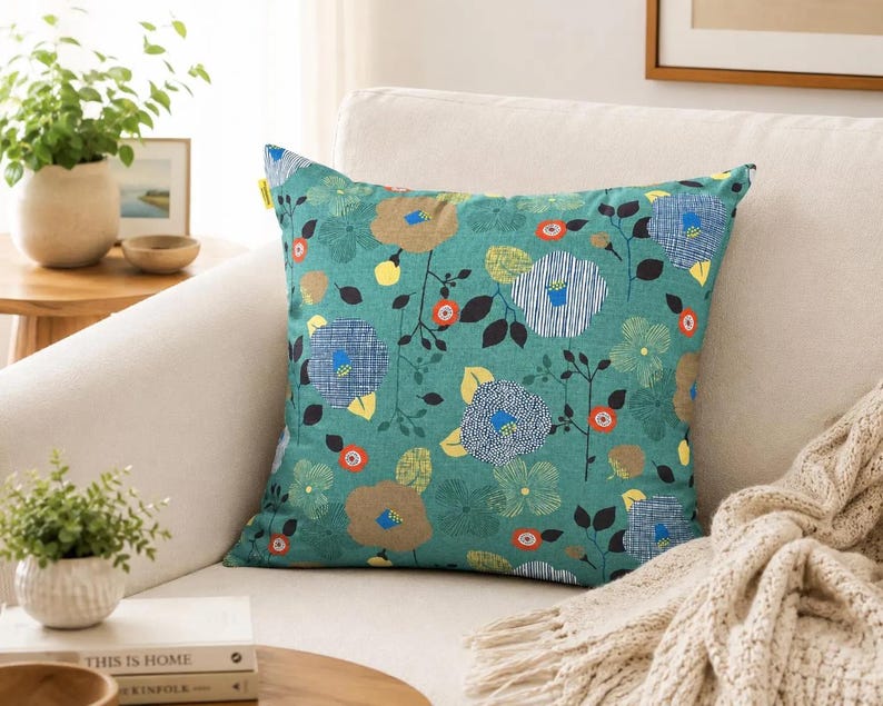 May include: A square throw pillow featuring a teal green background with a repeating pattern of stylized flowers in shades of blue, tan, yellow, and orange, accented by black stems and leaves. A small yellow tag is visible on the upper left side.