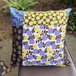 Peut inclure: Un coussin carr&eacute; pr&eacute;sentant un motif patchwork de fleurs. Les panneaux de tissu affichent des fleurs jaunes et violettes sur des fonds blancs, marron et violet clair, cr&eacute;ant un arrangement botanique multicolore sur toute la surface.
