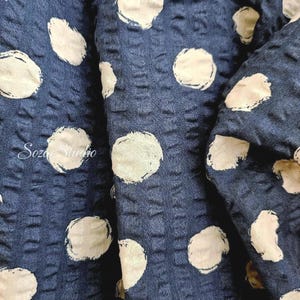 May include: Close-up of a navy blue fabric with a textured surface and large, off-white polka dots. The fabric is folded, revealing its depth and pattern. The text "Soza Studio" is visible in the lower left corner.