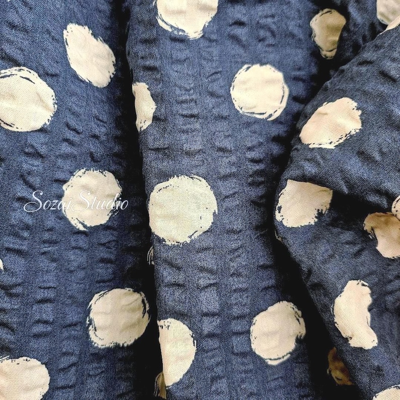 May include: Close-up of a navy blue fabric with a textured surface and large, off-white polka dots. The fabric is folded, revealing its depth and pattern. The text "Soza Studio" is visible in the lower left corner.