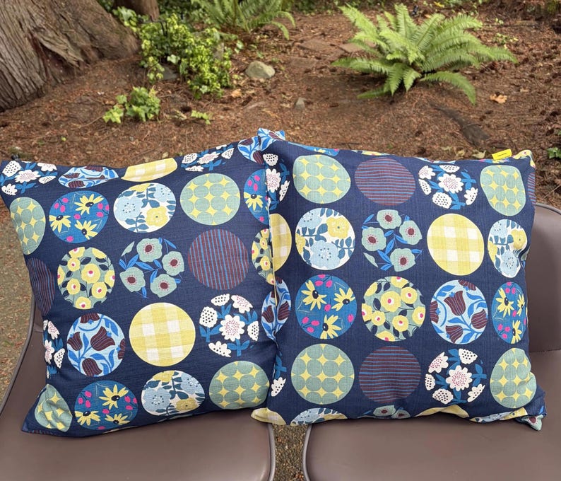 May include: Two square throw pillows featuring a dark navy blue background with a repeating pattern of circles. Each circle contains distinct designs including yellow gingham, blue and yellow floral prints, and geometric star or striped motifs.